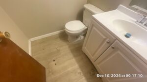 Bathroom Cleaning Before - Bio Decon Solutions Small bathroom with a white toilet beside a white vanity, beige walls, and a dirty, debris-strewn floor.