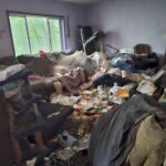 Dilapidated living room filled with trash, bags, and debris piled on the floor by a window outside greenery visible through the glass.