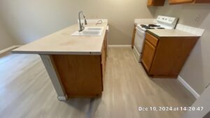 Kitchen Cleaning After 1 - Bio Decon Solutions Kitchen with an island and double-basin sink, chrome faucet, and white stove against light walls; wooden cabinets and beige countertops.
